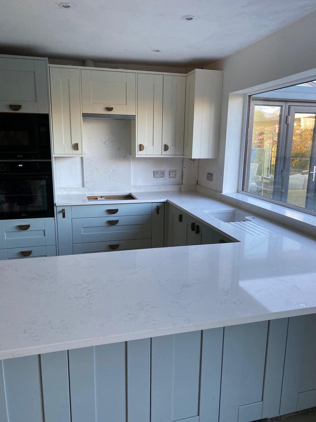 A fitted kitchen installation with light gray-blue base cabinets, cream upper cabinets, and white marble-effect quartz countertops with an island, integrated black oven on the left, and large windows overlooking the garden.