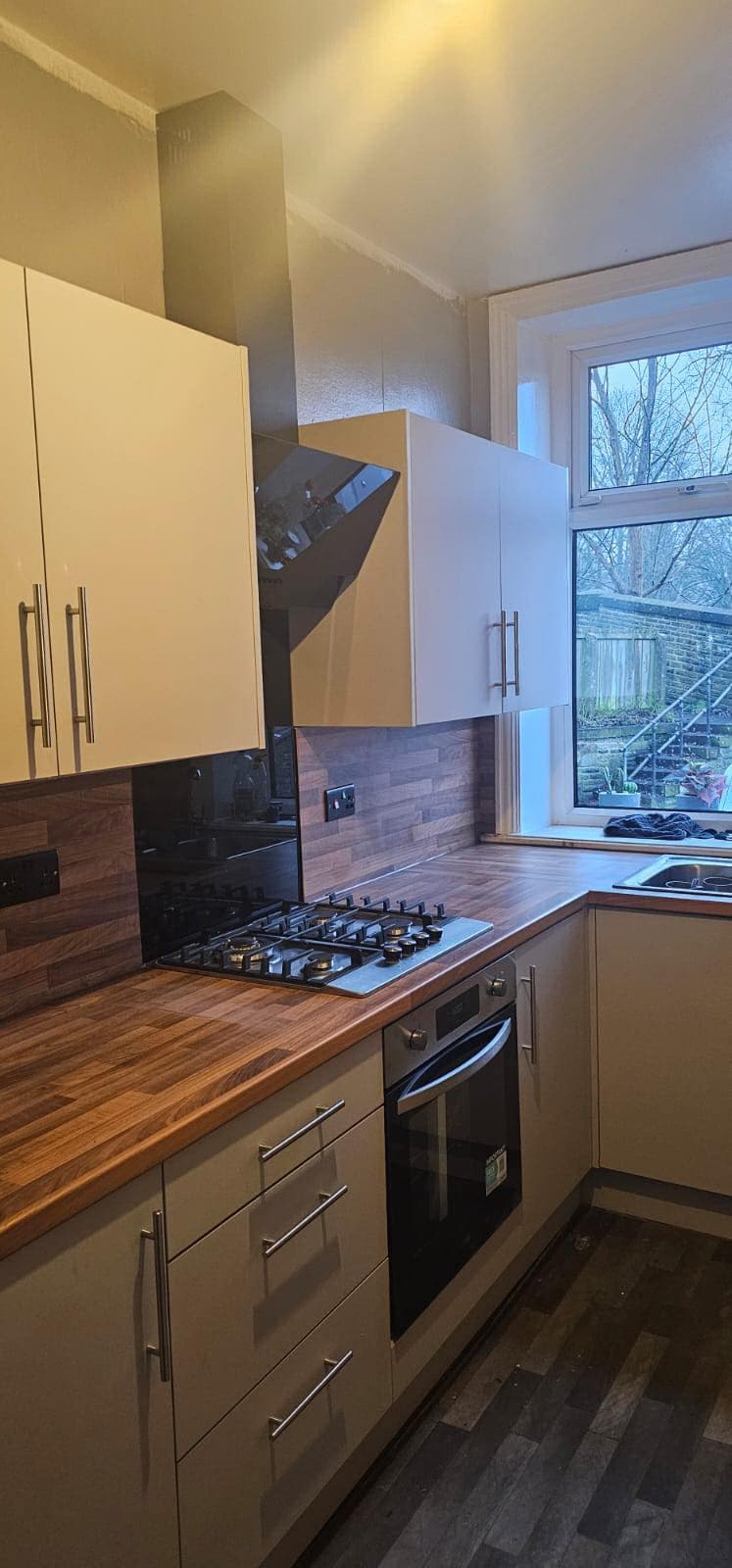 Modern cream and white kitchen with wooden worktops, black gas hob and oven, and wood-effect brick backsplash, featuring floor-to-ceiling cupboards and a corner window.
