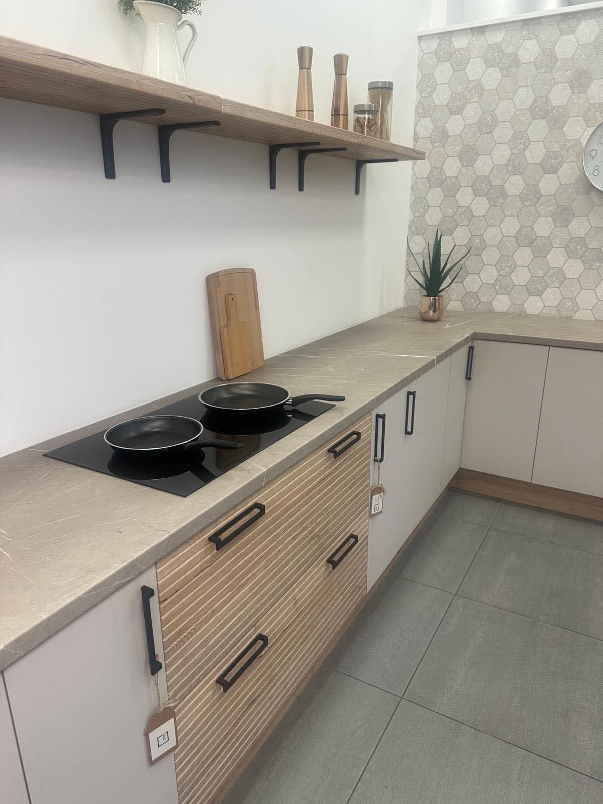 Installed modern kitchen featuring white and wood-grain cabinetry, beige countertops, hexagonal tile backsplash, induction cooktop, floating wooden shelves with black brackets, and grey tile flooring.