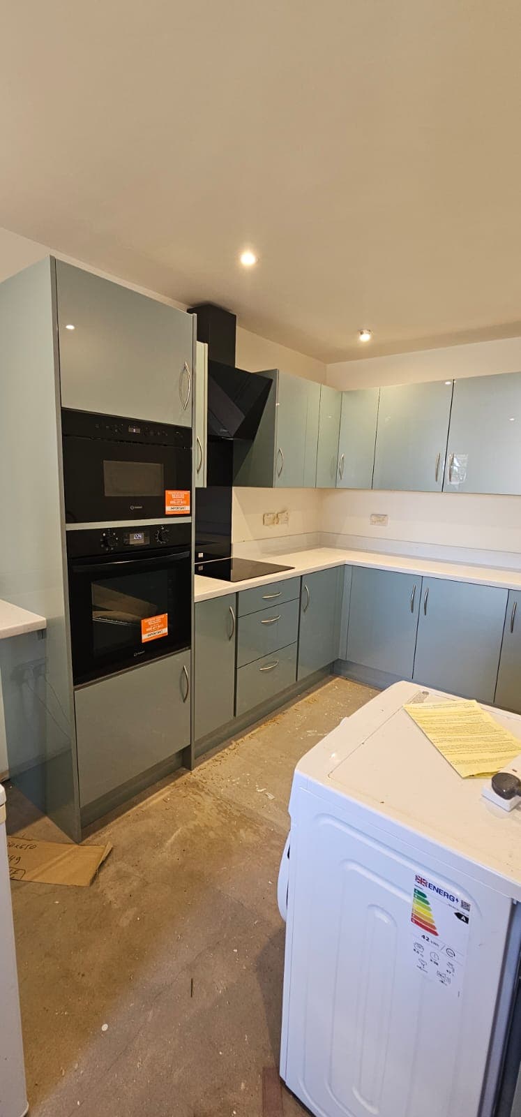 A newly installed modern kitchen with grey and white cabinetry, black built-in ovens, white countertops, and recessed lighting in the ceiling, still under construction with protective coverings on the floor and a washing machine visible in the foreground.