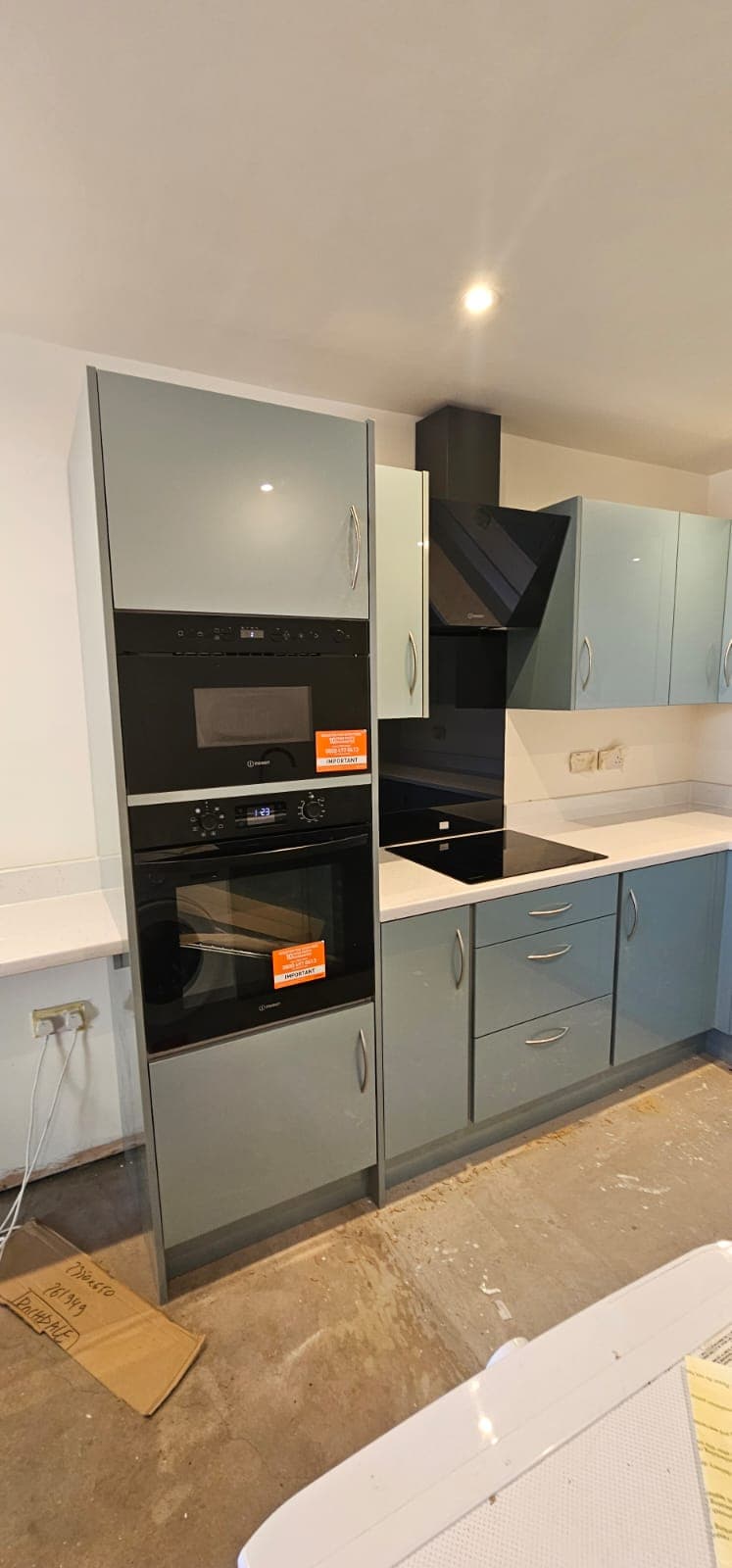Installed fitted kitchen with grey base cabinets, white and grey upper cabinets, black double ovens, black hob, white worktops, and ceiling lights in a newly built or renovated space.