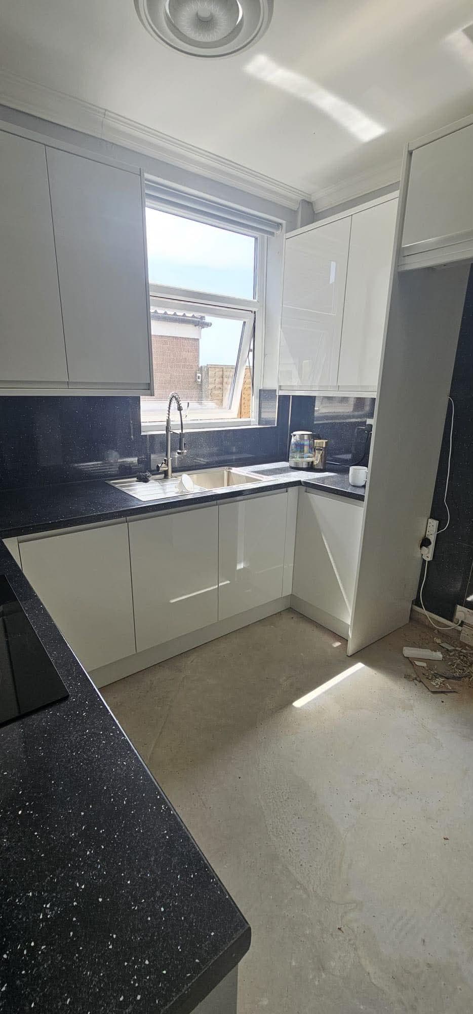 Installed kitchen with white gloss fitted cabinets, black speckled quartz worktops, stainless steel sink with tap, window with blinds, and recessed ceiling light.