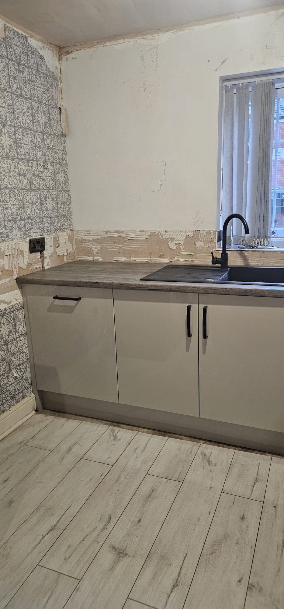 Galley cream kitchen with grey wood-look worktop, window view, patterned backsplash tile, and black sink.