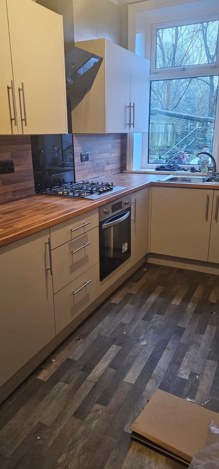 Cream shaker kitchen with walnut worktop, dark backsplash tile, integrated black oven, gas hob, and large window.