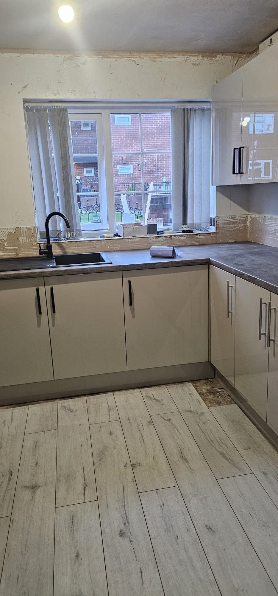 Cream painted u-shape kitchen with grey wood-look worktop, integrated black appliances, light wood flooring, and window.