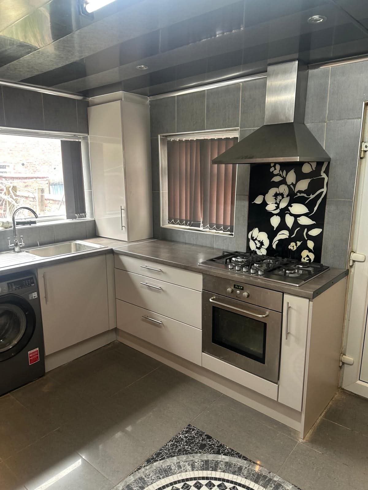 Cream cabinetry with grey stone worktop, dark grey tiled backsplash and decorative black floral splashback behind a gas range cooker, integrated washing machine visible in a corner unit kitchen.
