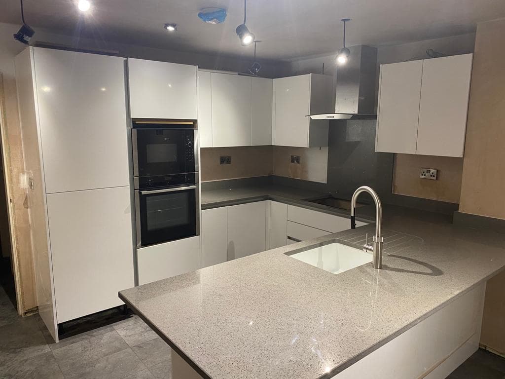 Modern white handleless kitchen with light grey quartz worktops, central island with sink, integrated black double oven, and pendant lighting above.