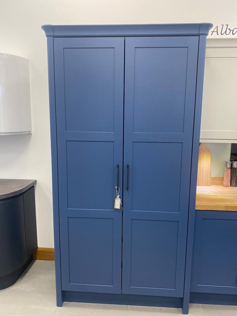 A tall navy blue painted kitchen cabinet with six recessed panels and dark handles, positioned against a cream wall in a showroom setting.