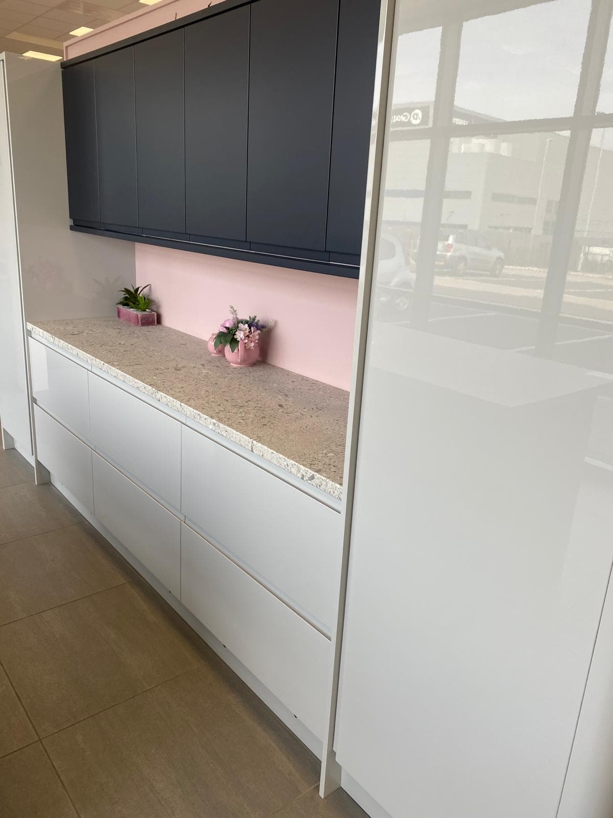 Two-tone kitchen with dark grey upper cabinets and white lower cabinets, featuring a speckled stone worktop and pink painted splashback.