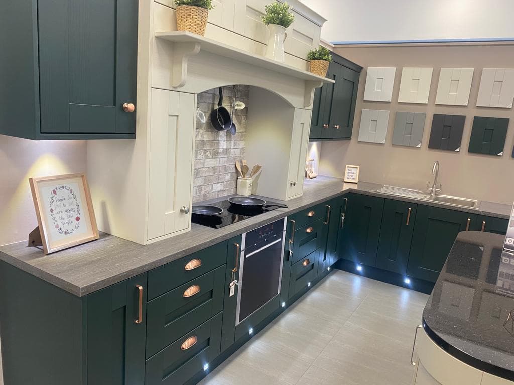 Two-tone shaker kitchen with dark forest green and cream cabinetry, grey stone worktops, and copper hardware, featuring an arched alcove with brick splashback and integrated lighting under the units.