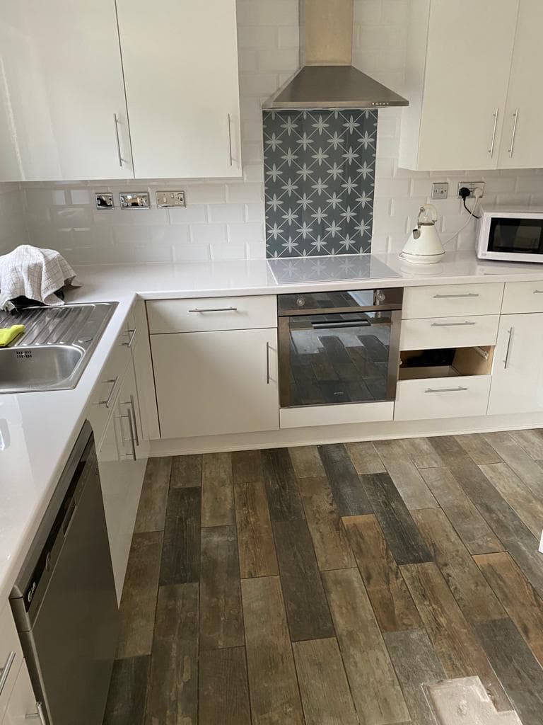 White in-frame kitchen with grey and white geometric patterned splashback, stainless steel range hood, and integrated oven against wood-effect flooring.