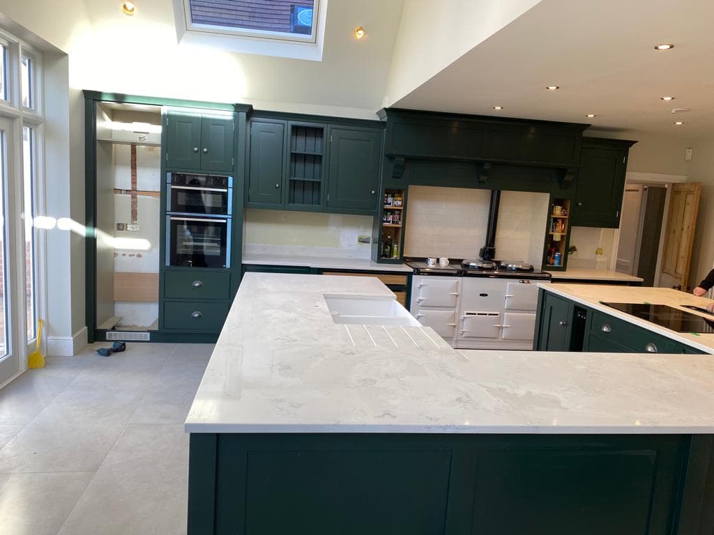 Modern kitchen with dark forest-green painted cabinetry, white marble worktops, and a large central island with seating, featuring integrated appliances and pendant lighting.