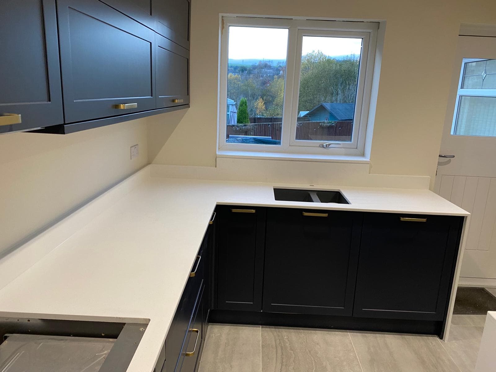 Modern kitchen with dark grey and black cabinetry, white quartz worktops, and brass handles, featuring a U-shaped layout with an island and large windows overlooking rural countryside.