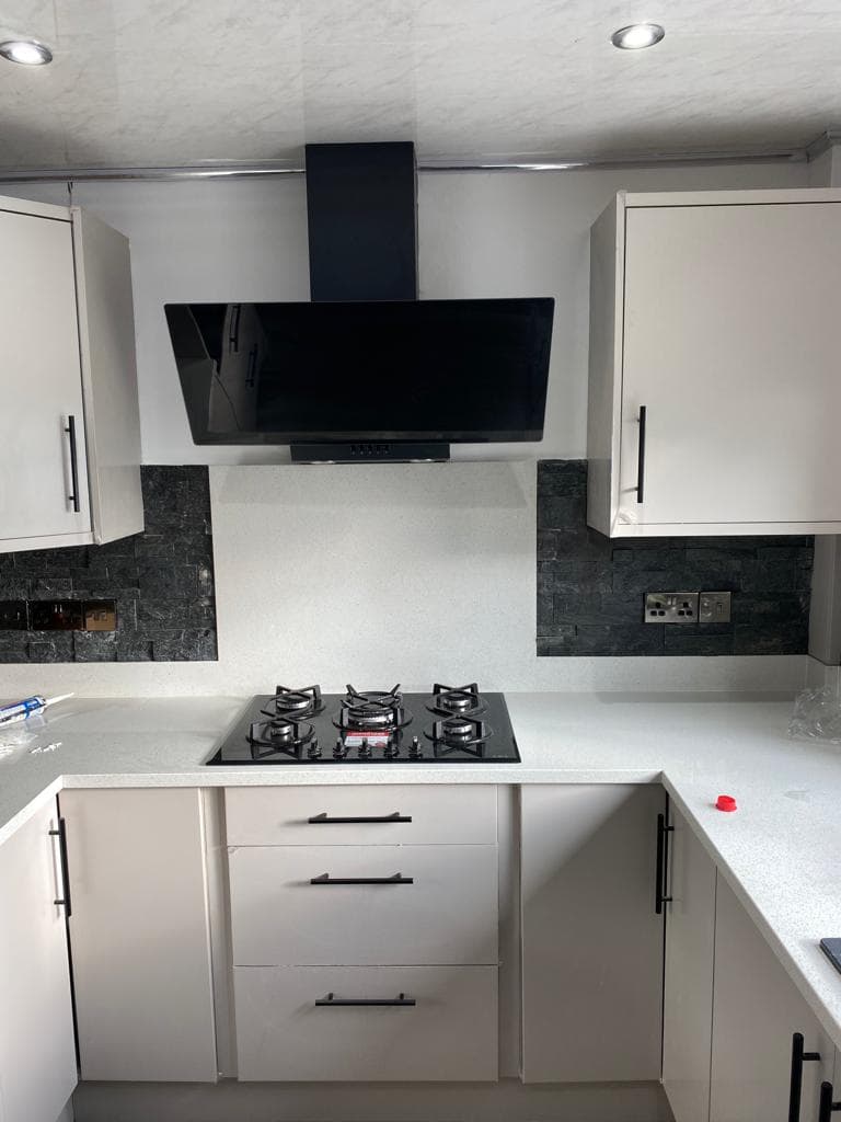 Modern kitchen with cream shaker-style cabinets, dark slate splashback, white quartz worktop, black gas hob, and black angled extractor hood.