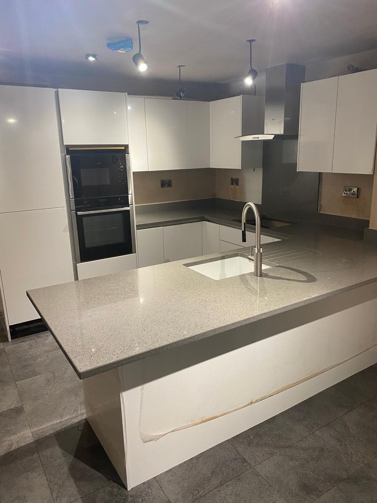 Modern kitchen with white gloss cabinetry, grey quartz worktops, central island with integrated sink, double built-in ovens, and pendant lighting.