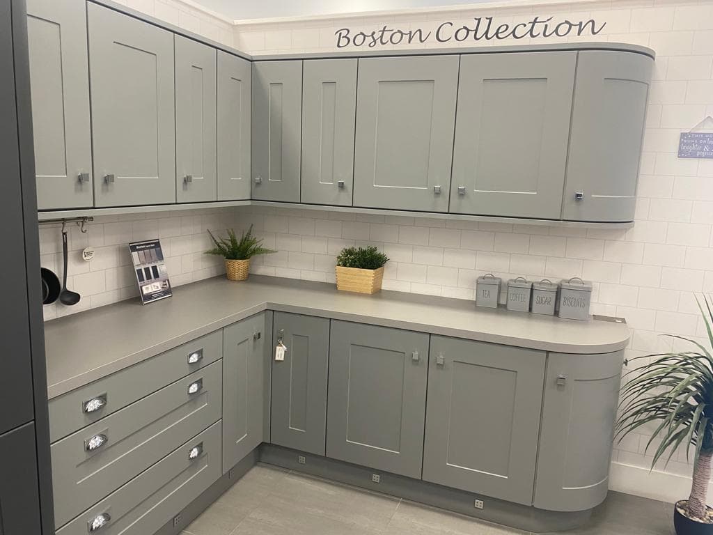 L-shaped kitchen with sage green painted cabinetry, white metro tile splashback, and light grey quartz worktops, featuring curved corner units and chrome handles.