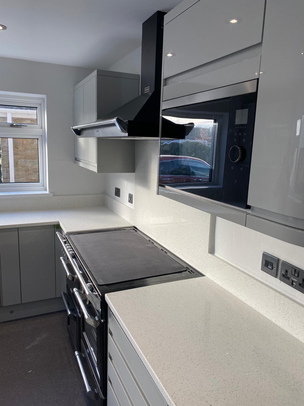 Modern kitchen with light grey and white cabinetry, white quartz worktops, integrated black double oven, black cooker hood, and stainless steel range cooker in an L-shaped layout.