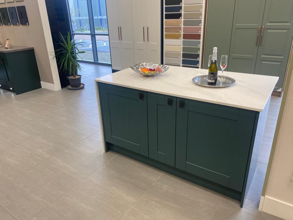 Modern kitchen with a dark sage-green island featuring a cream stone worktop, white cabinetry in the background, and large glass doors opening to a balcony.