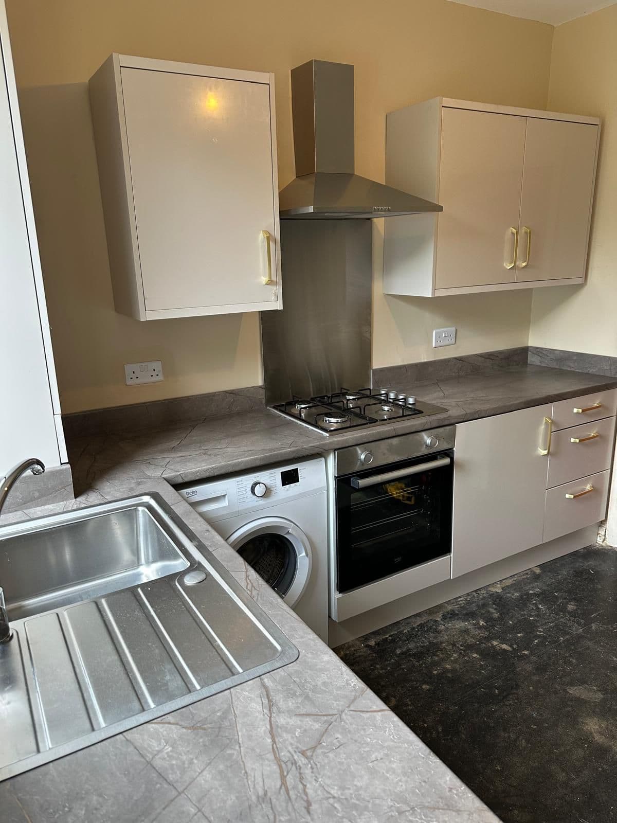 Newly installed modern kitchen with cream-coloured cabinets, dark grey stone countertops, stainless steel sink, integrated washing machine, black built-in oven and hob with extractor hood, and brass hardware handles.