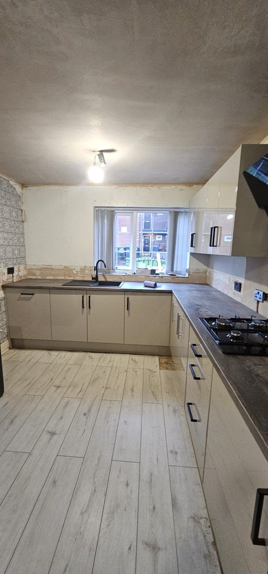Newly installed kitchen with cream-colored cabinets, dark worktops, gas hob, wood-effect flooring, and window overlooking a street.
