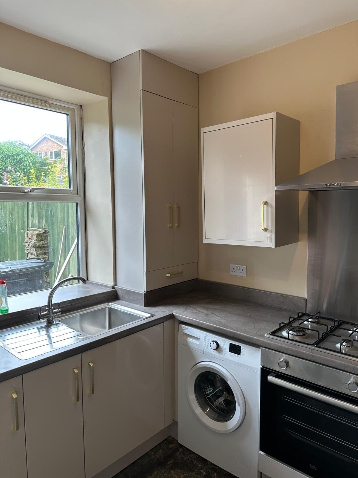 Installed kitchen with cream and taupe cabinetry, dark grey countertops, stainless steel appliances, white washing machine, gas hob, and window overlooking garden.