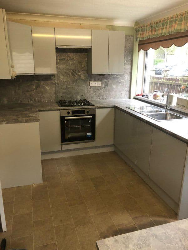 Installed fitted kitchen with cream-coloured units, dark marble-effect backsplash, black oven and gas hob, stainless steel sink and windows with decorative valance.