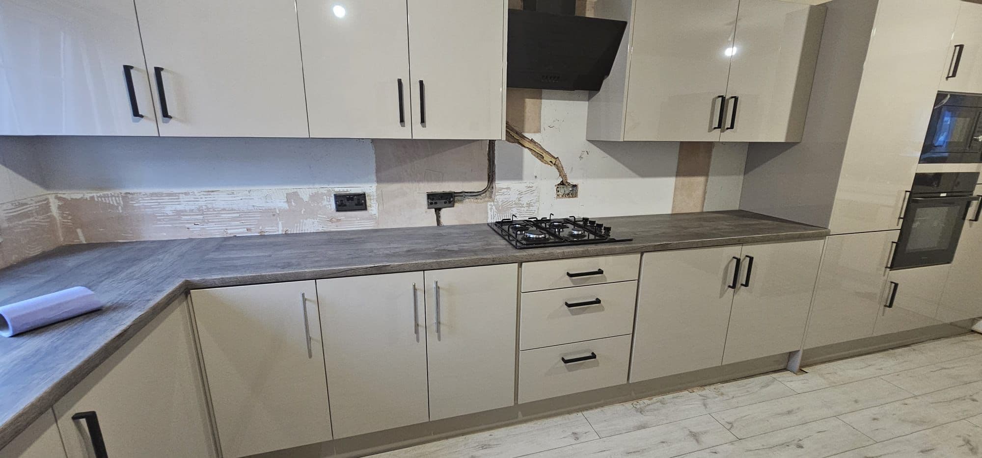 Installed fitted kitchen with cream-coloured units, dark grey worktops, built-in gas hob, overhead cabinets with black handles, and integrated built-in oven on the right side.