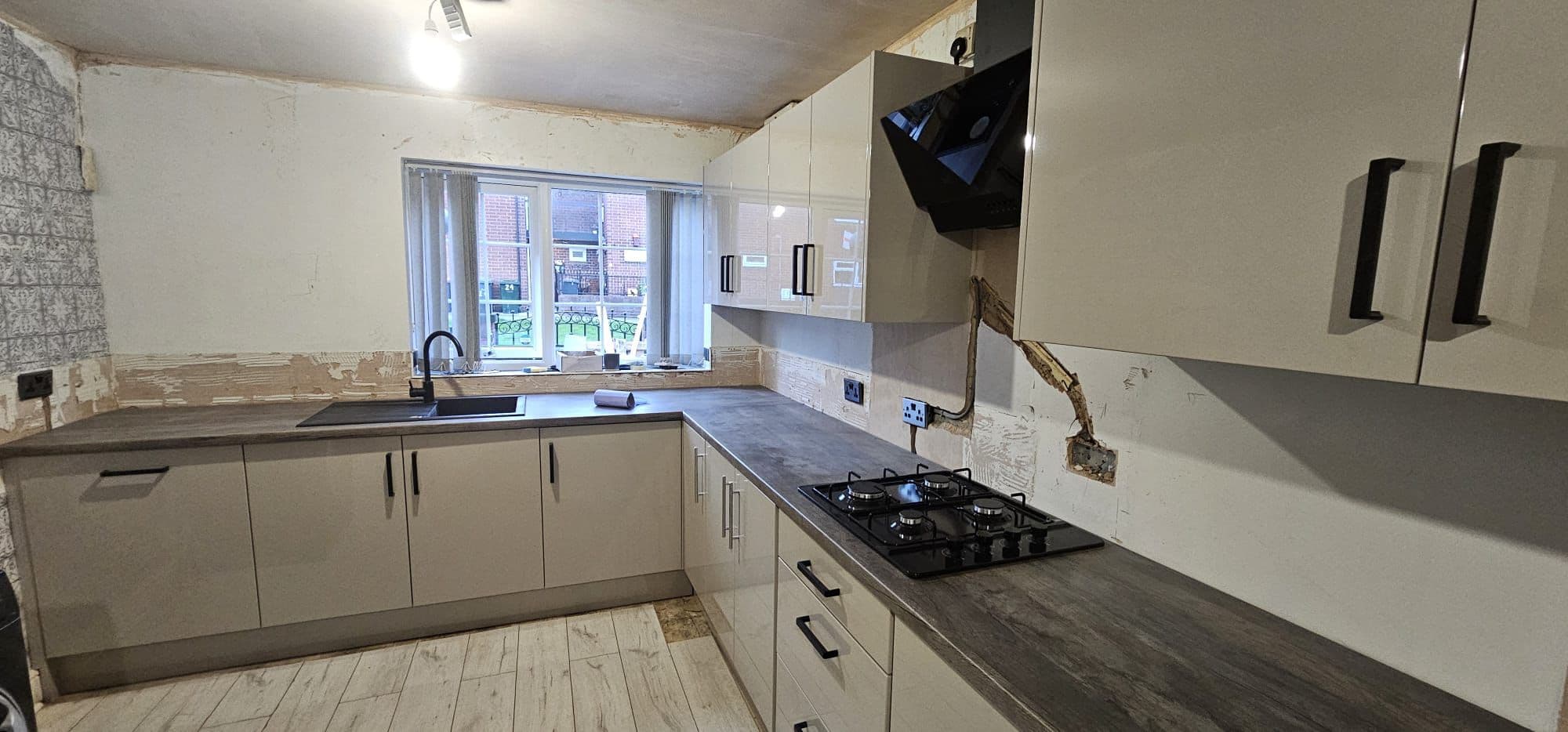 Installed fitted kitchen with cream-colored cabinetry, dark grey countertops, black gas hob, integrated appliances, and brick pattern backsplash tiling.